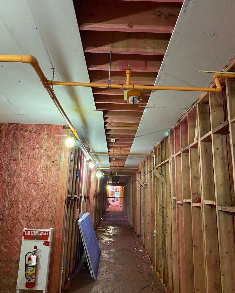 Wood-frame corridor with temporary sprinkler piping overhead and a fire extinguisher station